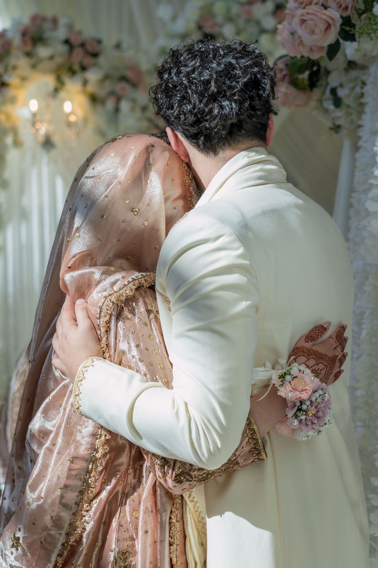 Bride and groom embracing at wedding ceremony, bride in gold embroidered dress with veil, groom in white suit.