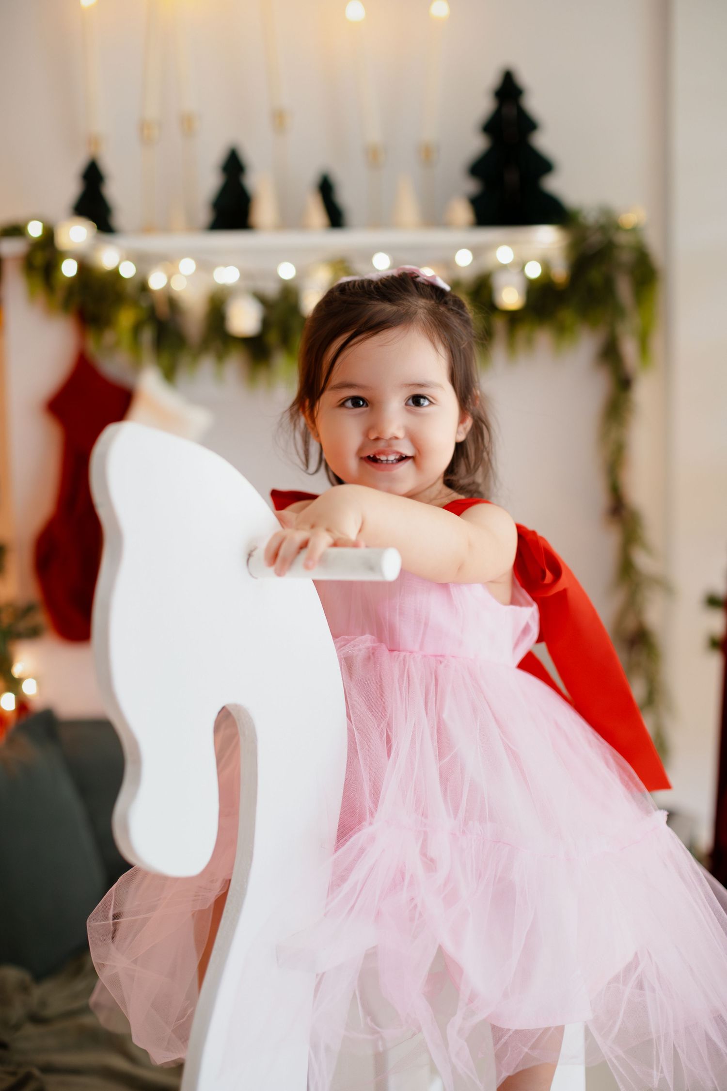 Smiling toddler girl in pink tutu dress riding white rocking horse near Christmas fireplace with lights.
