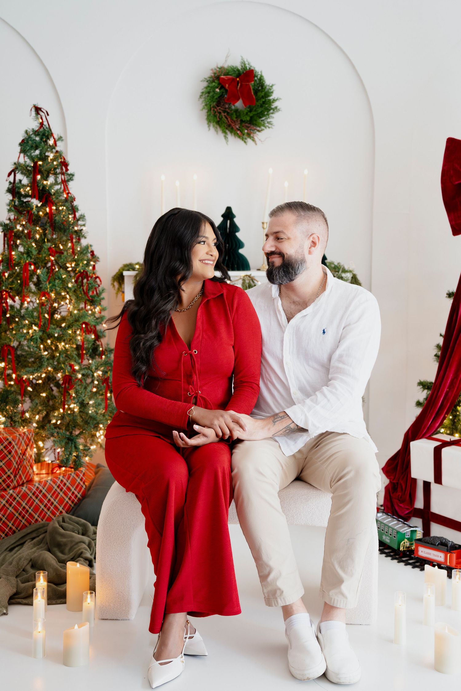 Couple in red and white outfits sitting together by a decorated Christmas tree with candles and wreaths.