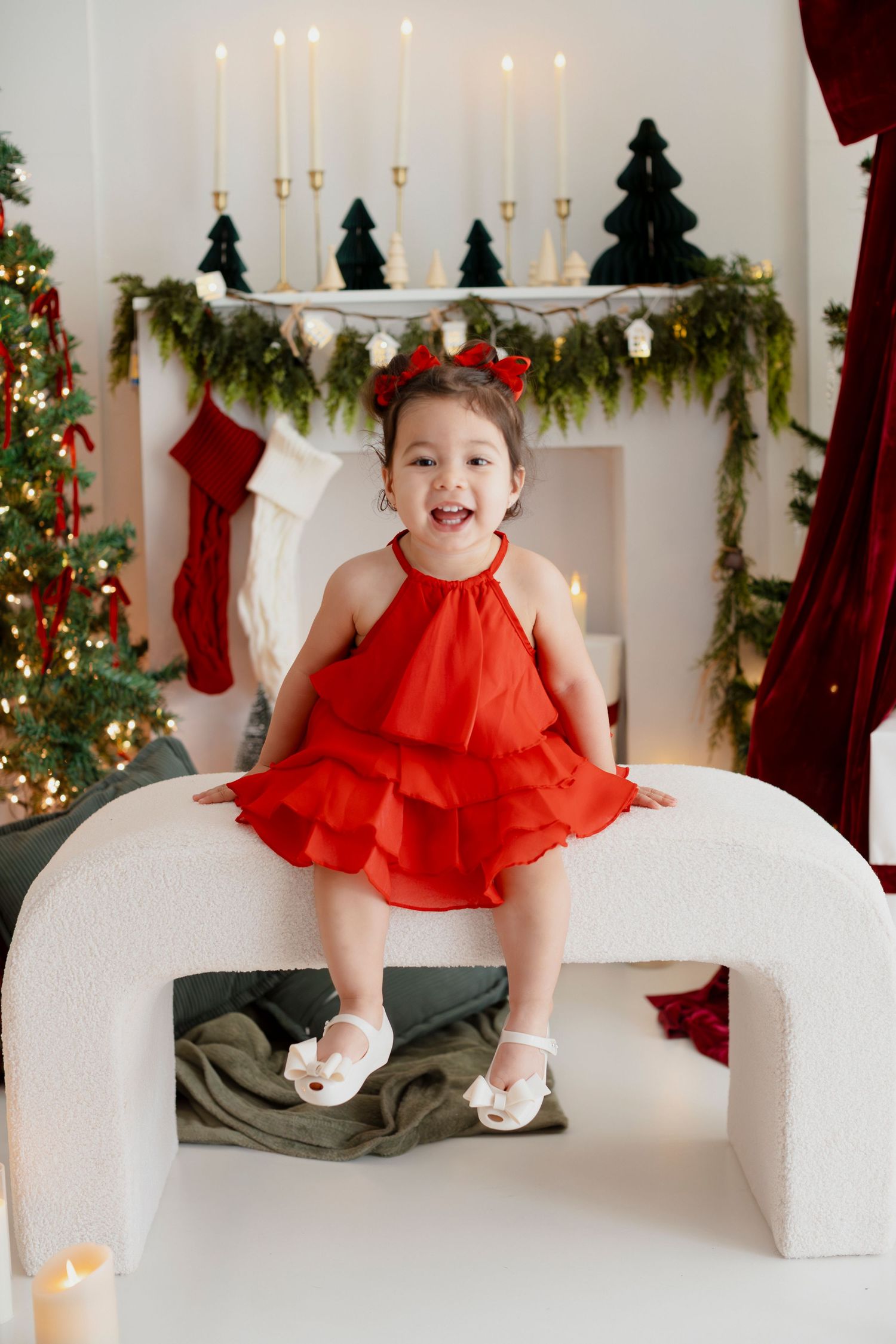 Smiling toddler girl in red ruffled dress and bow sits on white arch decor in festive Christmas room.