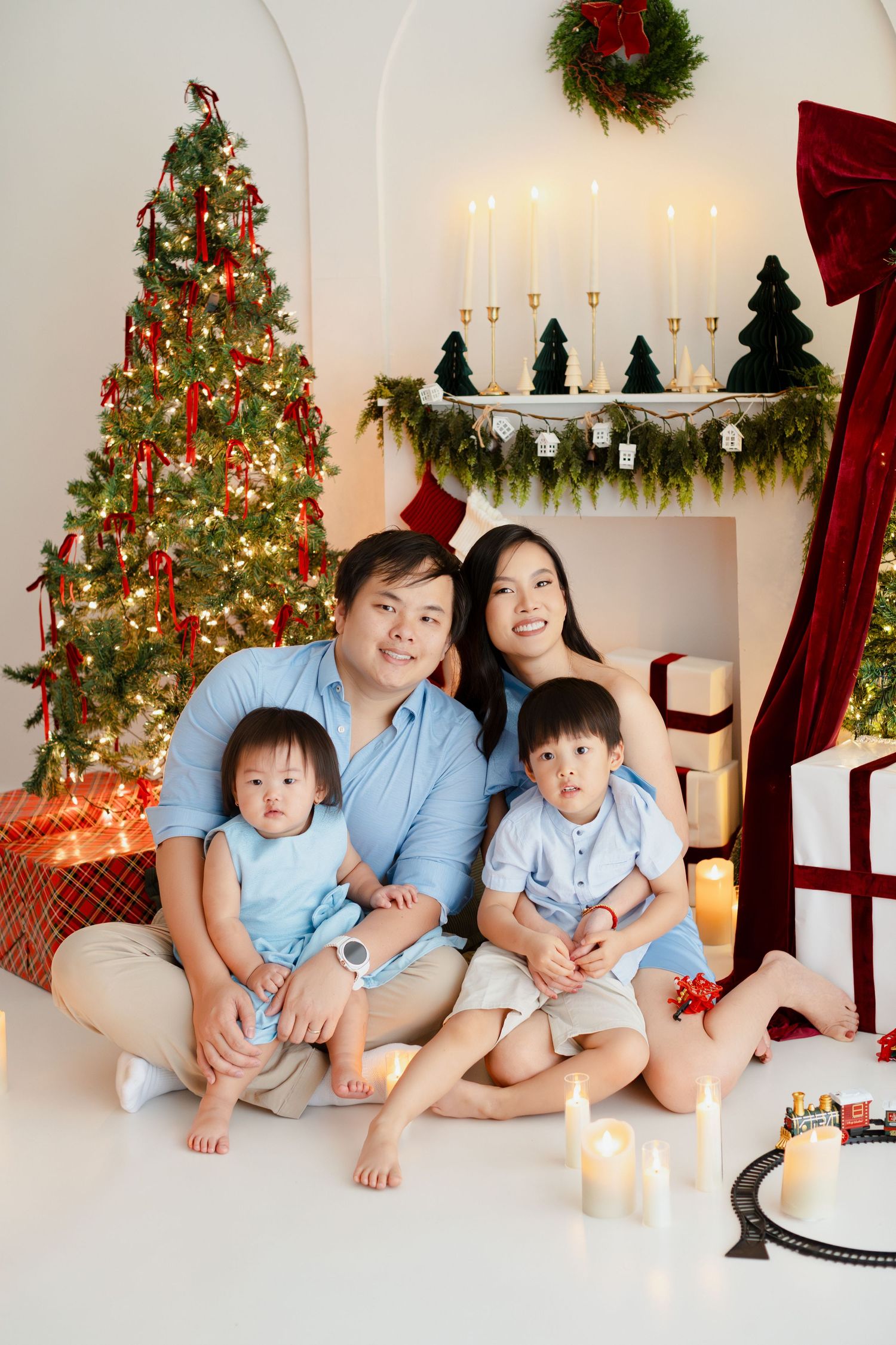 Asian family of four poses together in matching blue outfits by a decorated Christmas tree with gifts and candles.