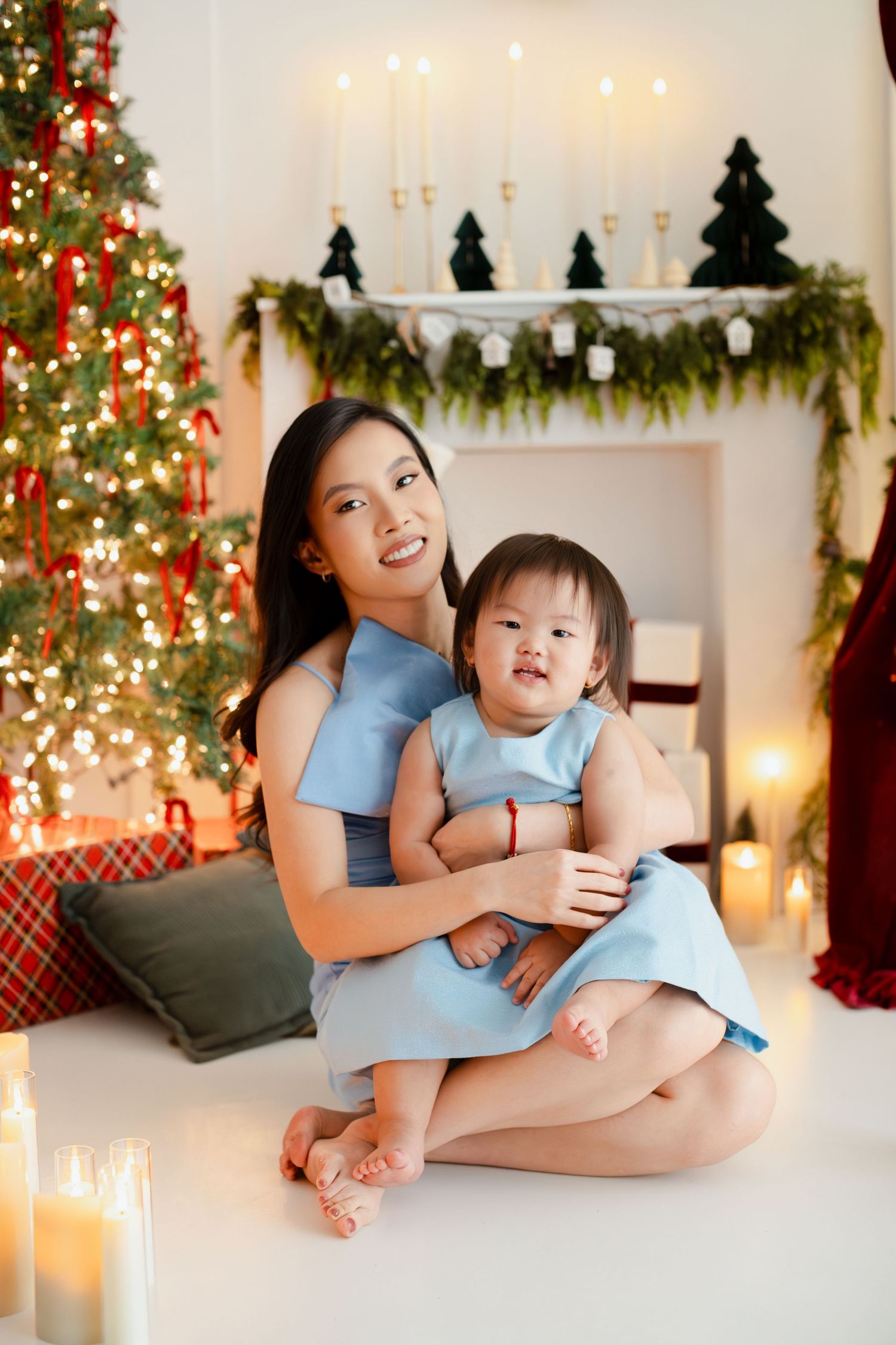 Asian mother and baby daughter in matching blue dresses pose by Christmas tree and fireplace with candles.