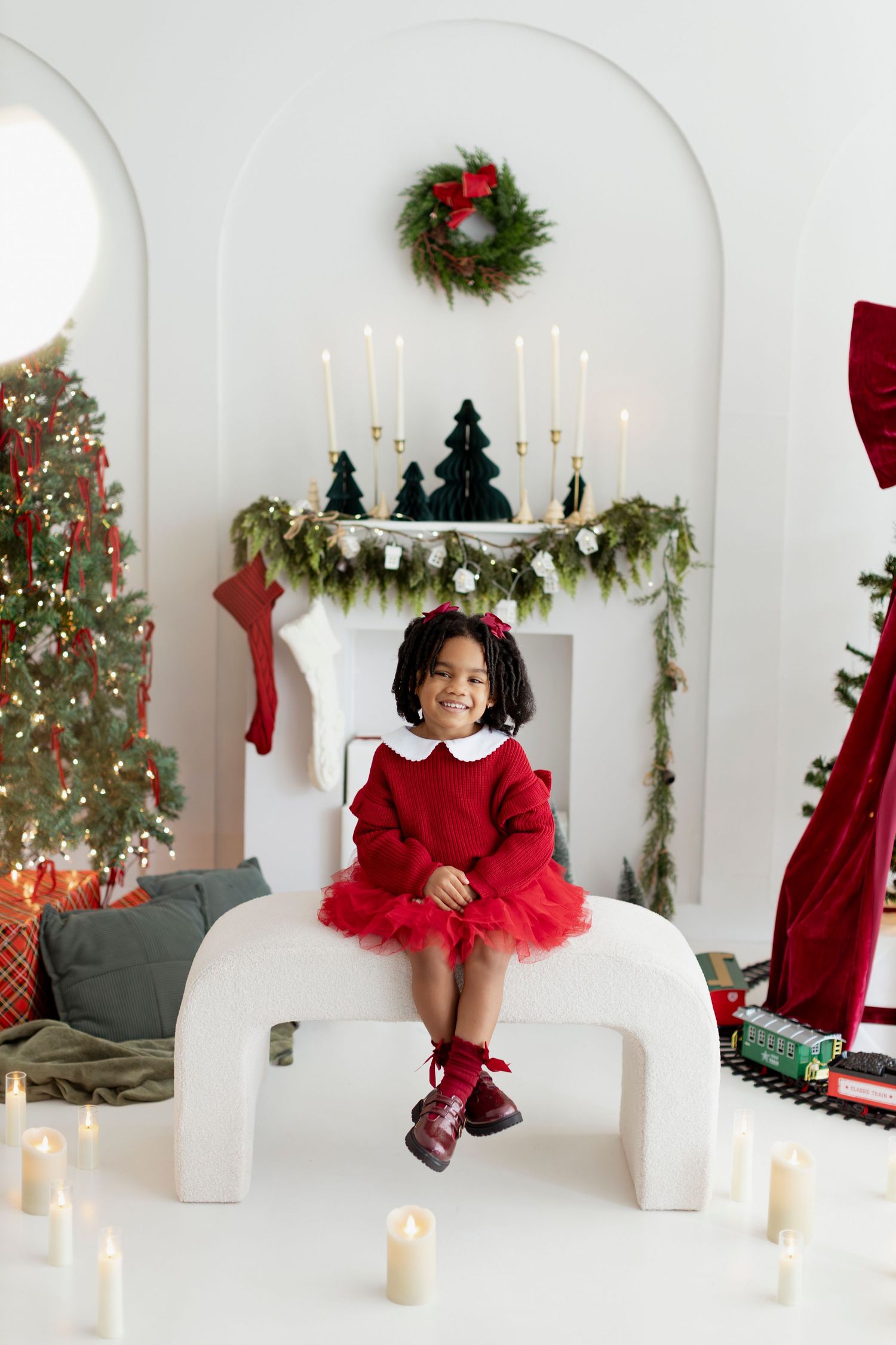 Young girl in red tutu dress sits on white bench amid Christmas decorations, candles, and festive wreath backdrop.