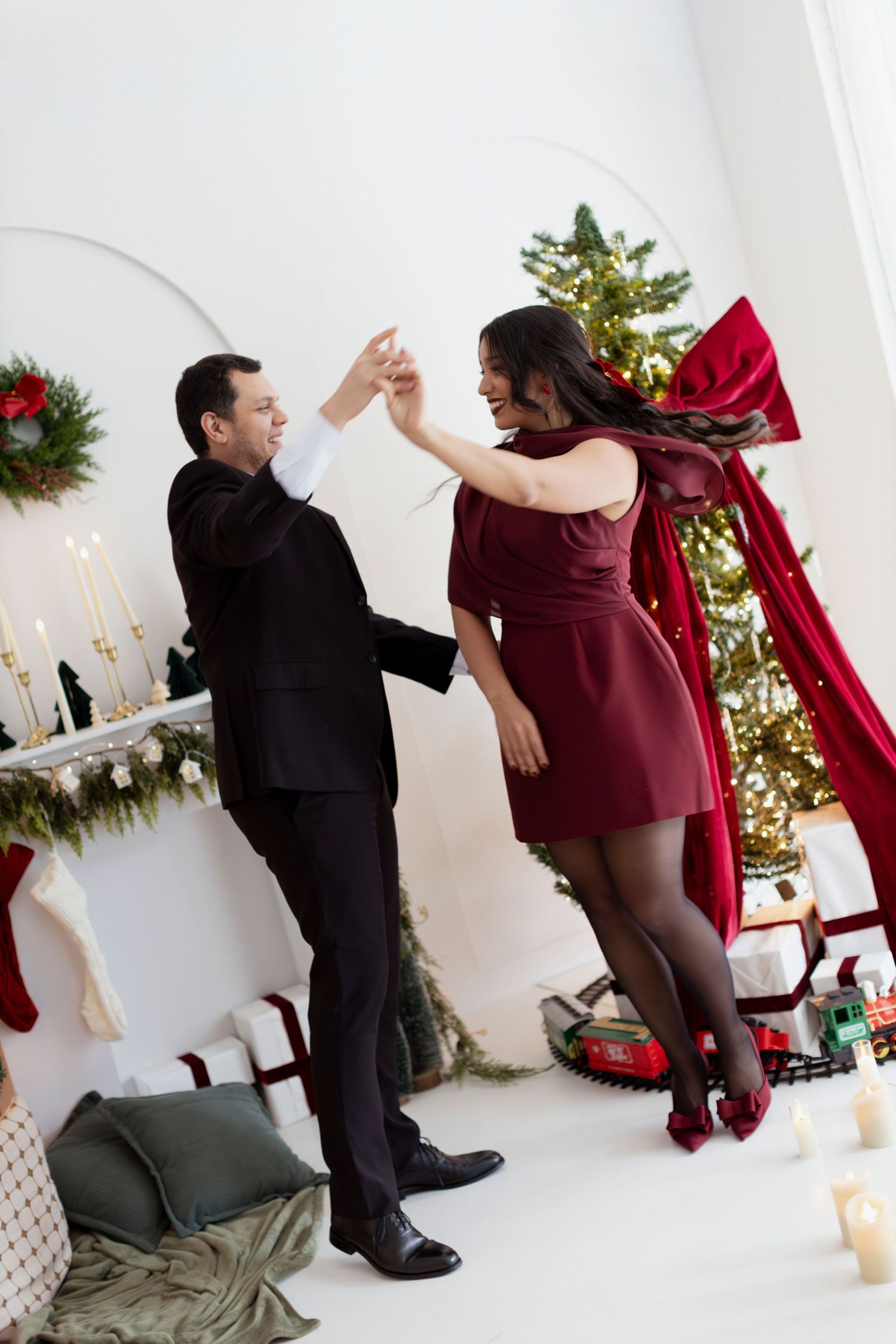 Couple dancing together in festive Christmas decorated room, woman in burgundy dress, man in black suit near decorated tree.