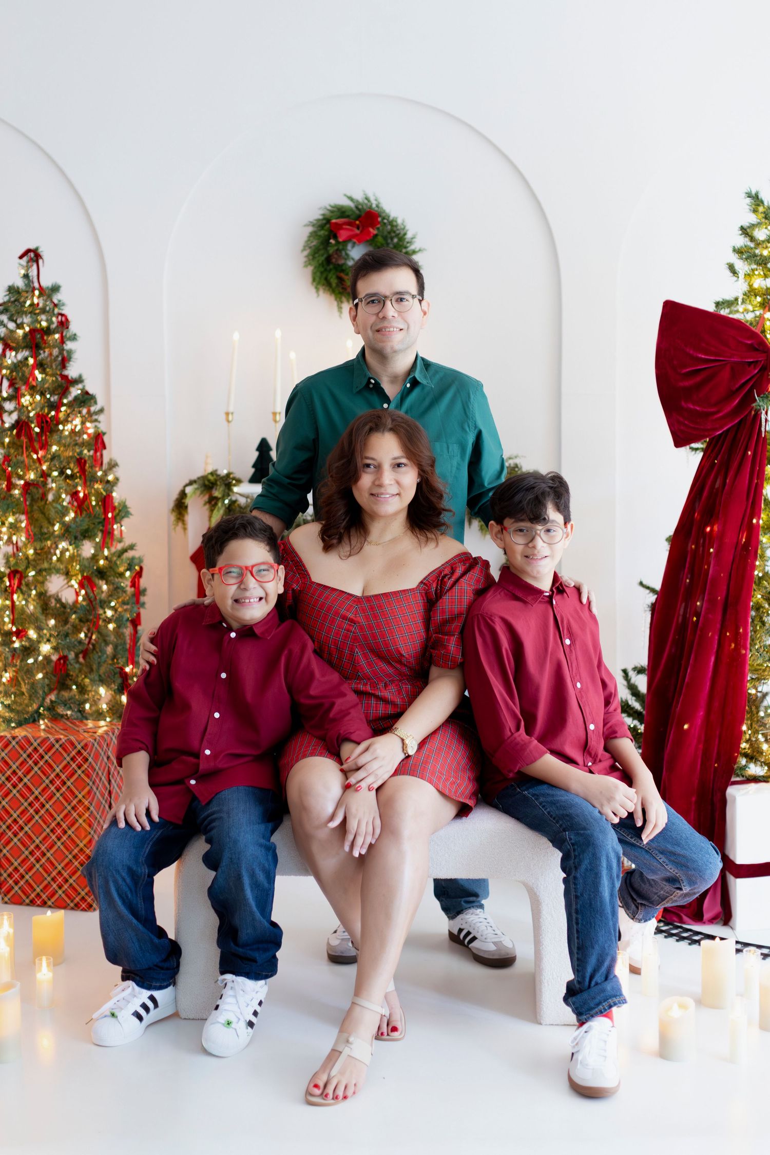 Family of four posing for a Christmas photo with decorated trees, candles, and holiday decor in a white studio setting.