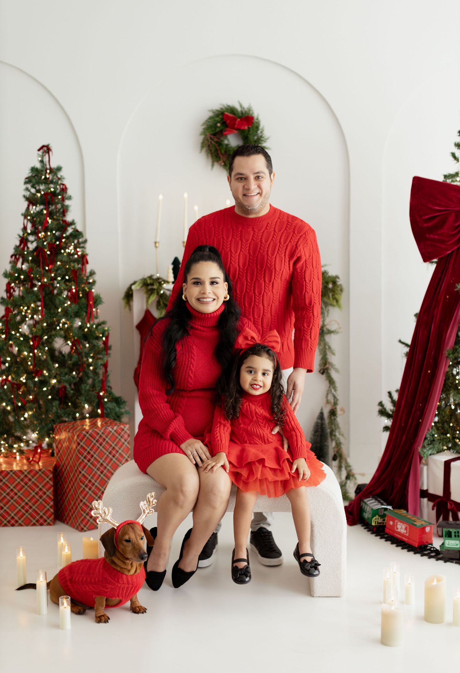 Family of three in matching red outfits pose with dachshund in Christmas sweater before decorated holiday tree.