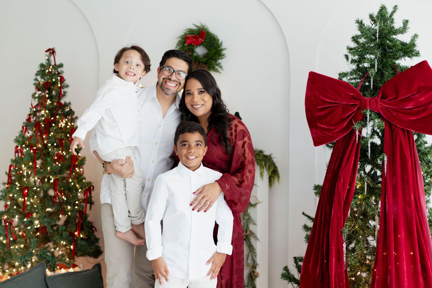 Family of four poses by decorated Christmas trees, wearing festive white and red outfits, smiling warmly indoors.