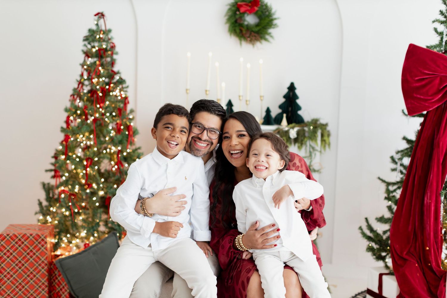 Family of four smiling together in front of a decorated Christmas tree, wearing white and red holiday outfits.