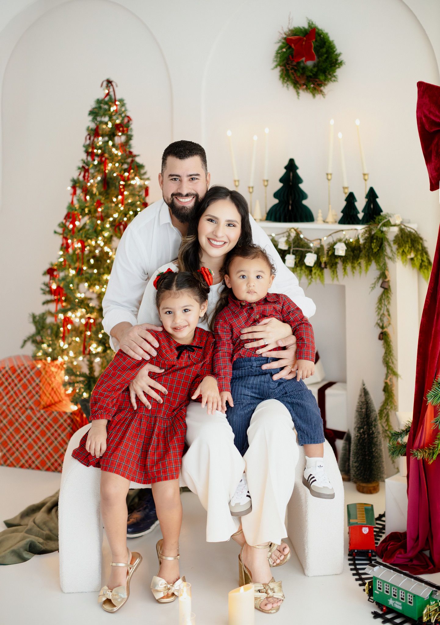 Family of four in matching red outfits posing together in a festive Christmas decorated room with tree and fireplace.