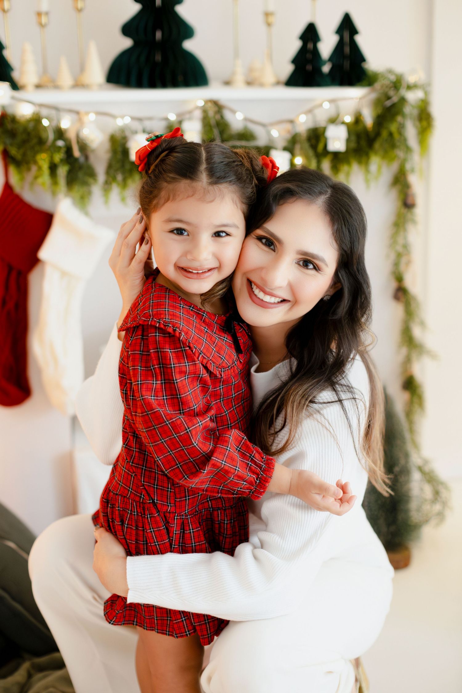 A mother and young daughter in festive Christmas outfits smile together in front of a holiday-decorated fireplace mantel.