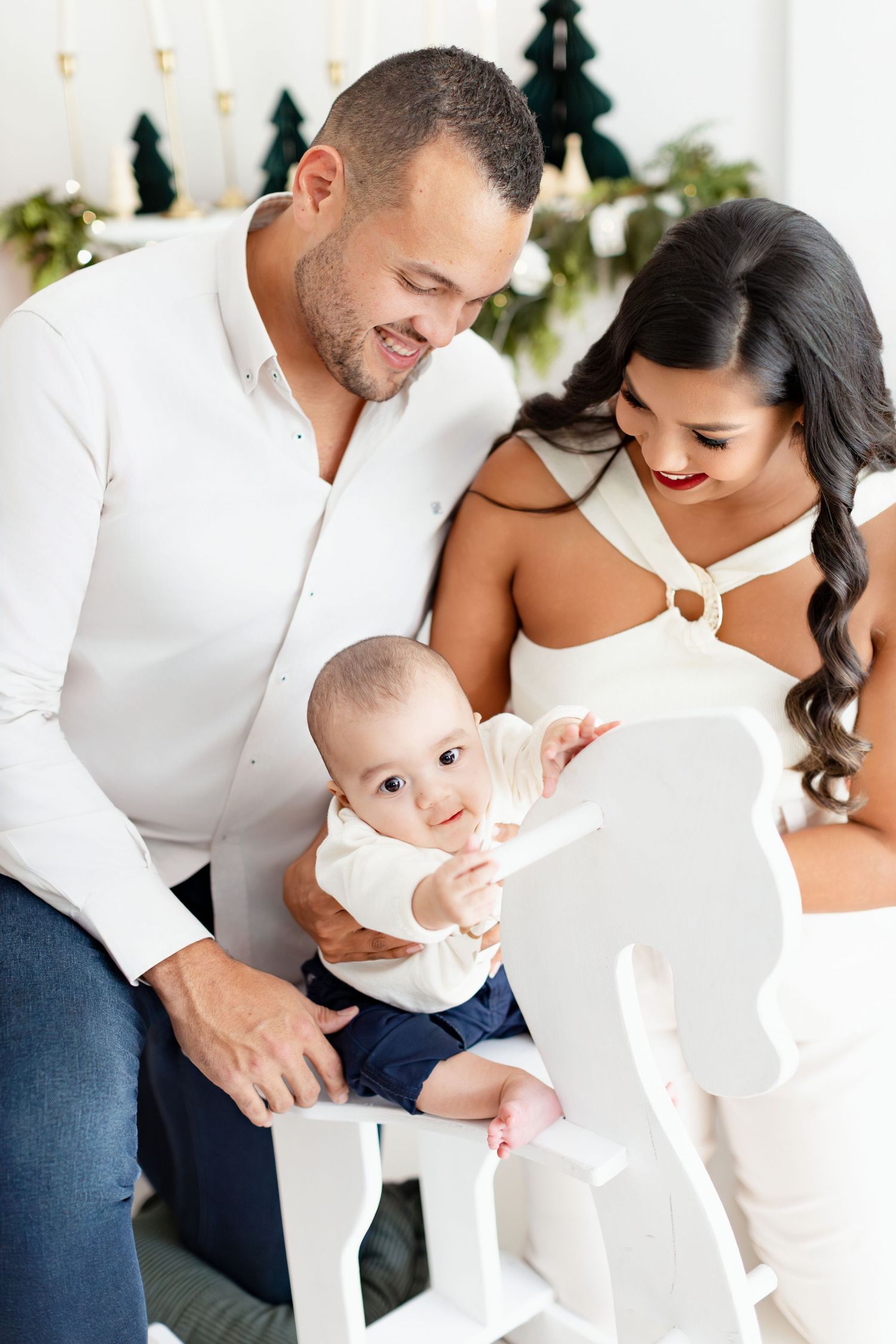Happy family with baby on white rocking horse, Christmas decor background, parents in white outfits smiling.