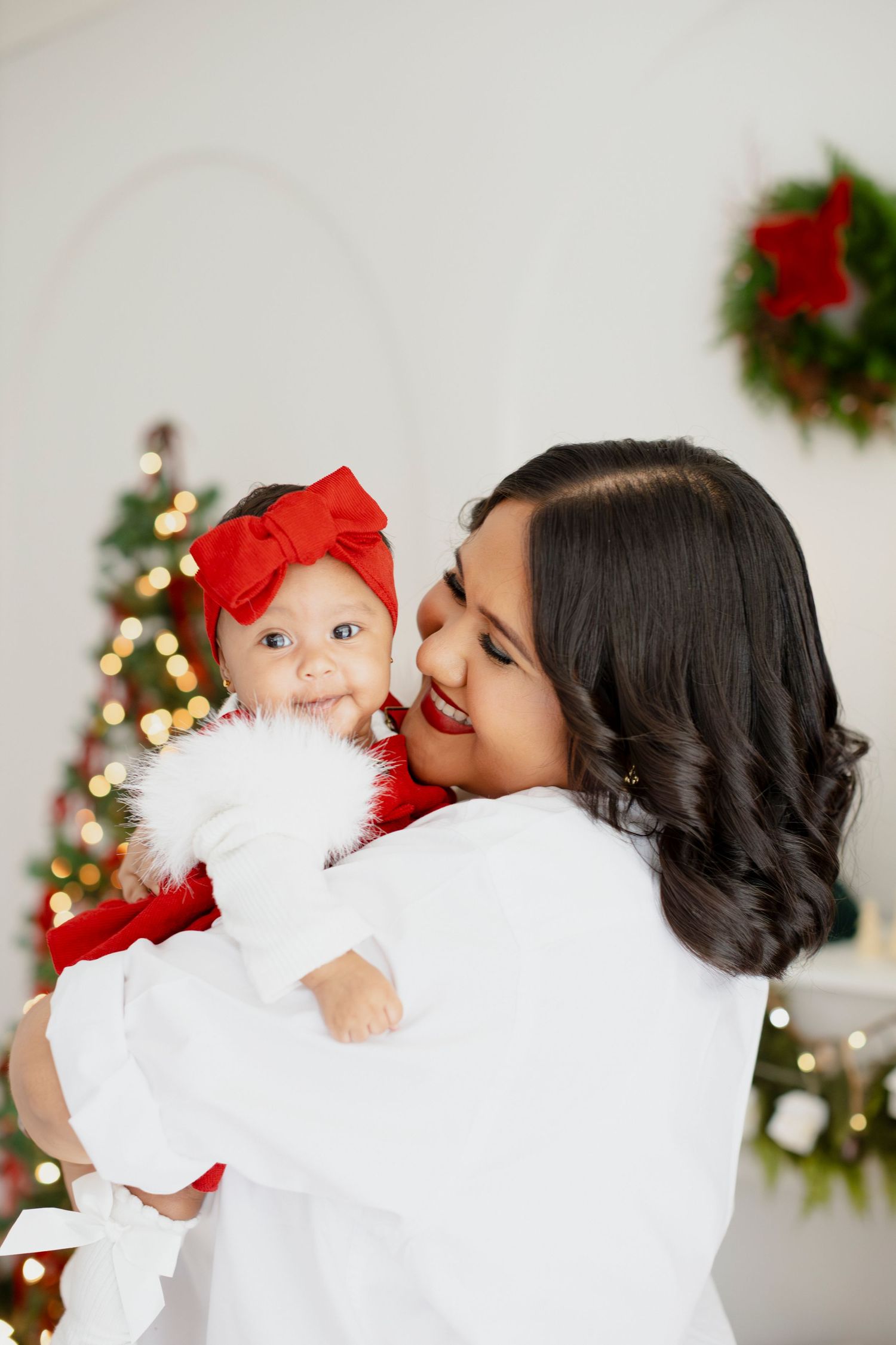 A mother in white holds her smiling baby in a Santa outfit with red bow near a decorated Christmas tree.