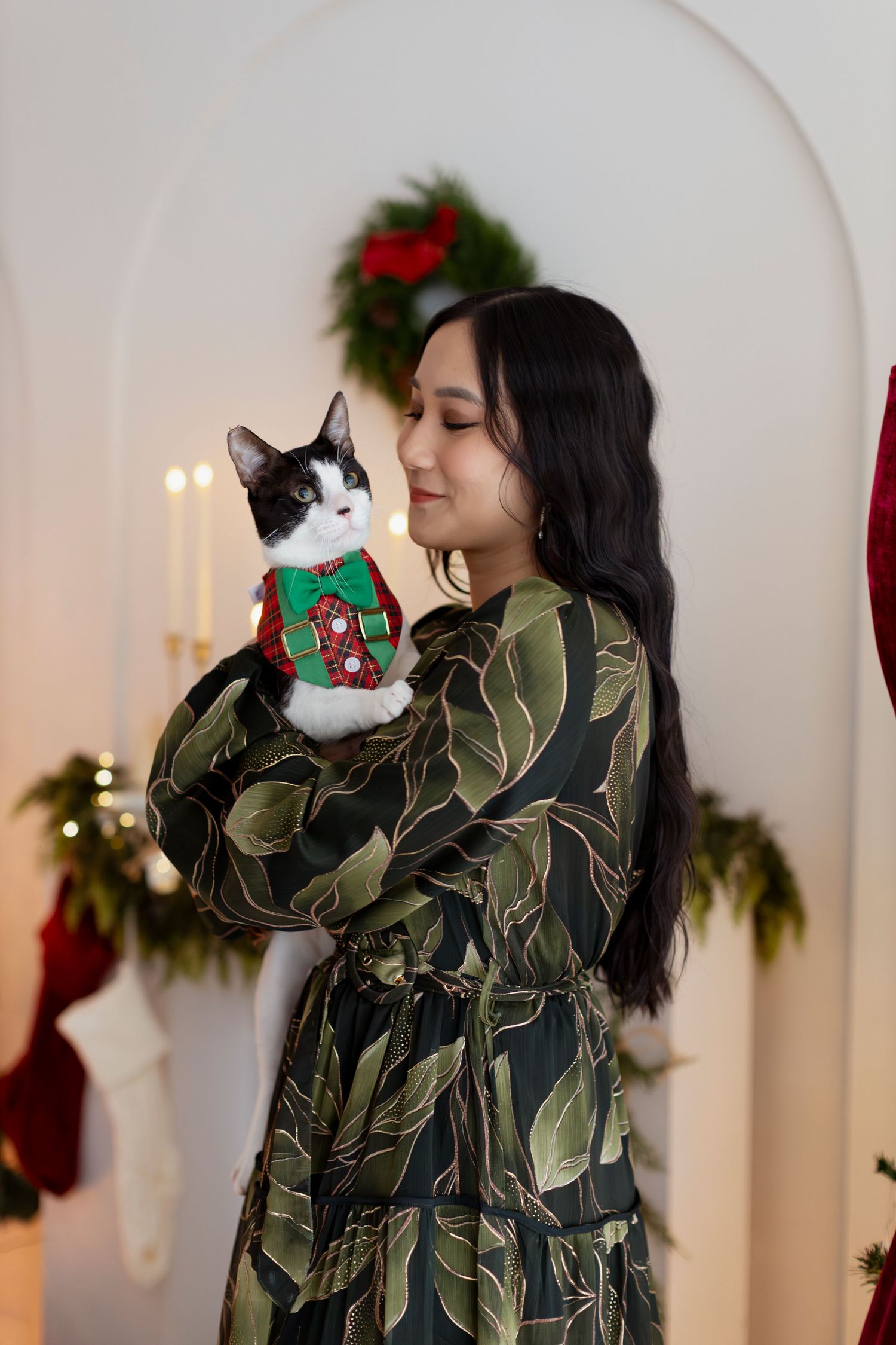 Woman in green floral dress holding a black and white cat in a Christmas sweater near holiday decorations.