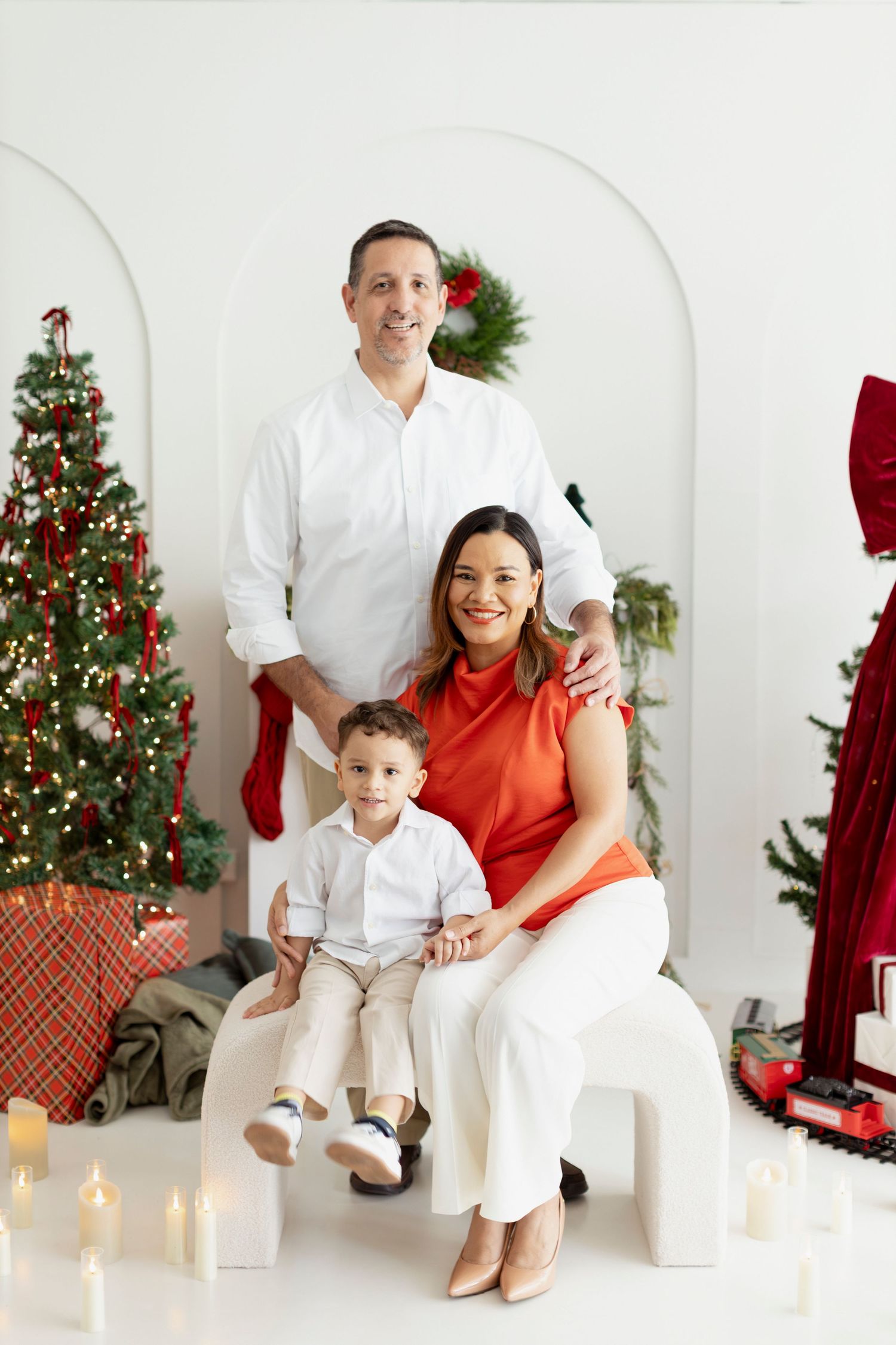Family of three posing for a Christmas portrait with decorated tree, candles, and holiday gifts in a white studio.
