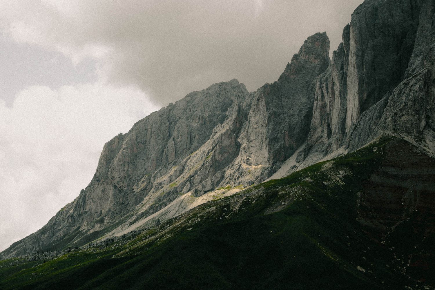 Dramatic rocky mountain peaks with steep gray cliffs rise above lush green slopes under a cloudy overcast sky.