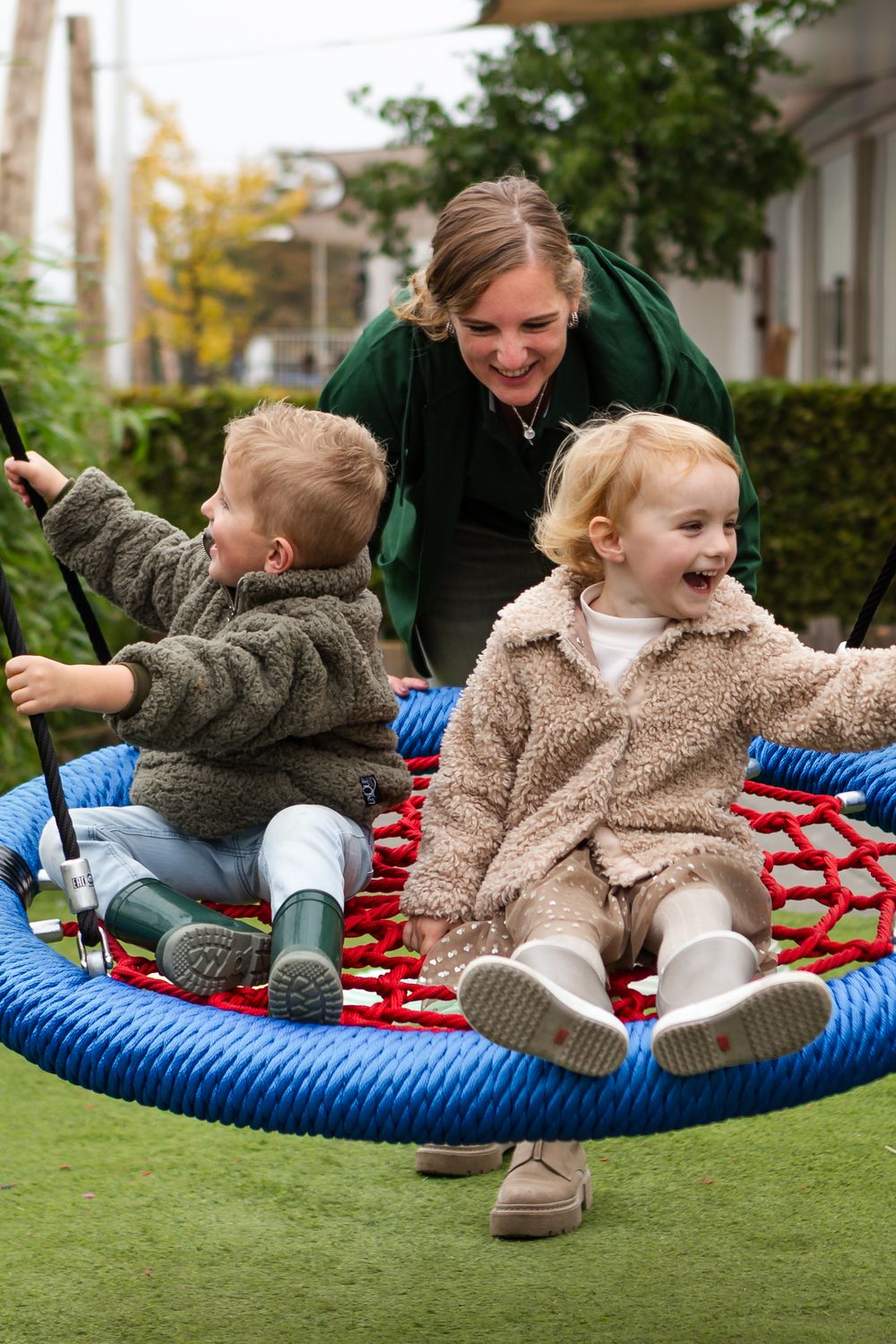 Children and adult enjoying playtime on a blue nest swing in a backyard garden setting.