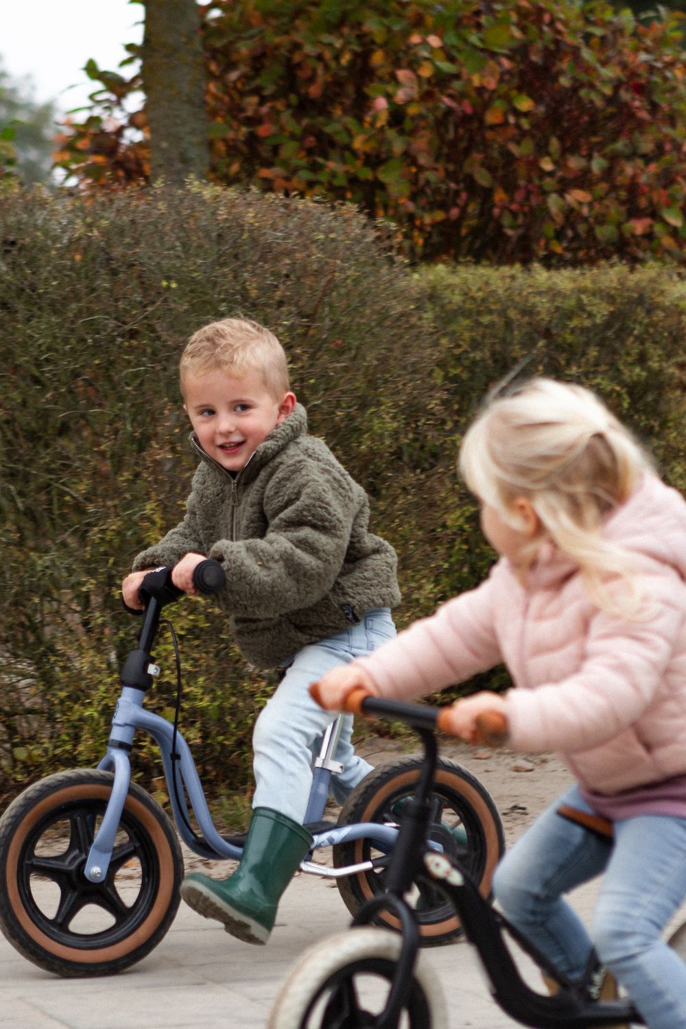 Two children riding balance bikes collide playfully on an autumn path surrounded by colorful fall foliage.