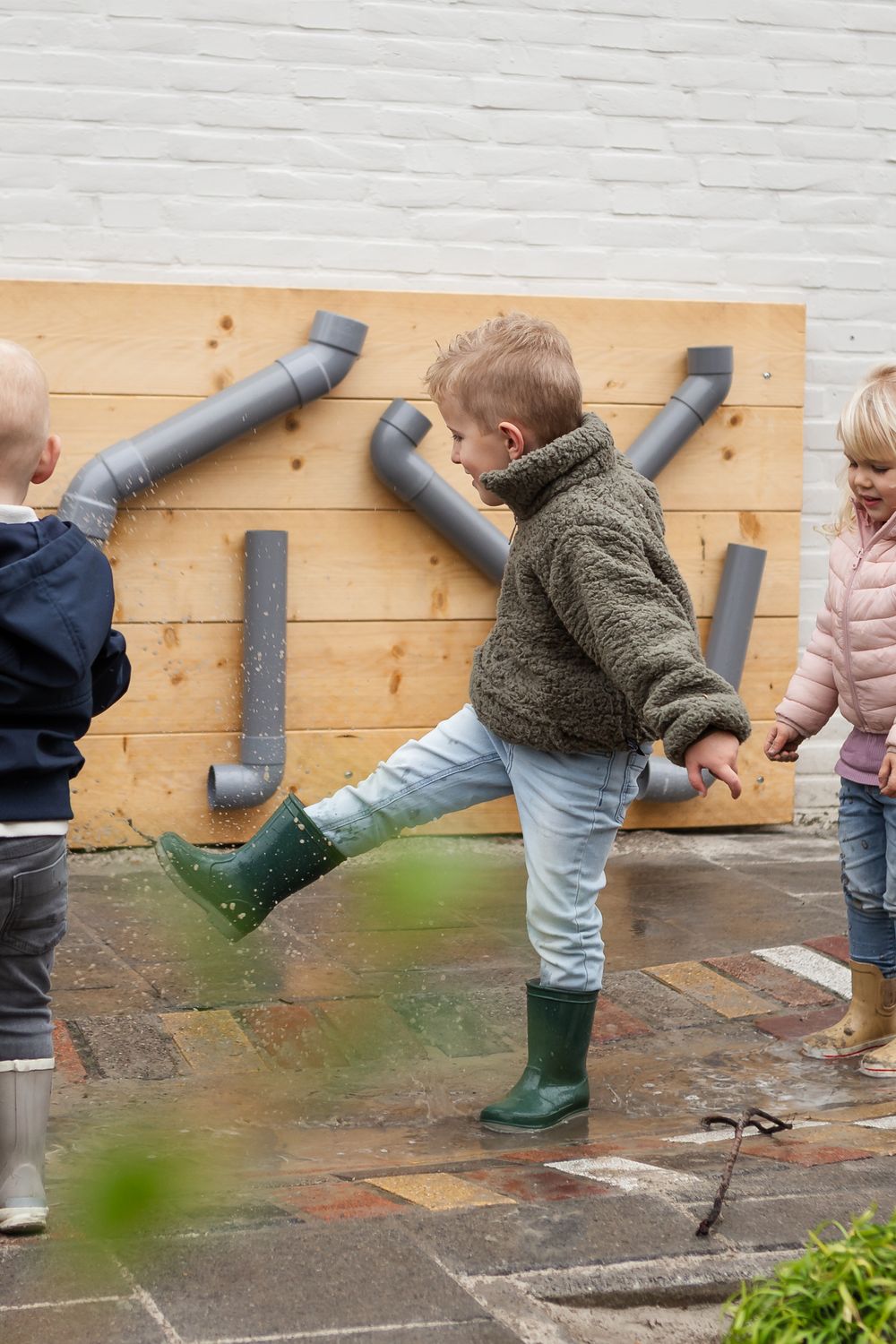 Children in rain boots explore a drainpipe during outdoor play, watching water flow through the pipe.