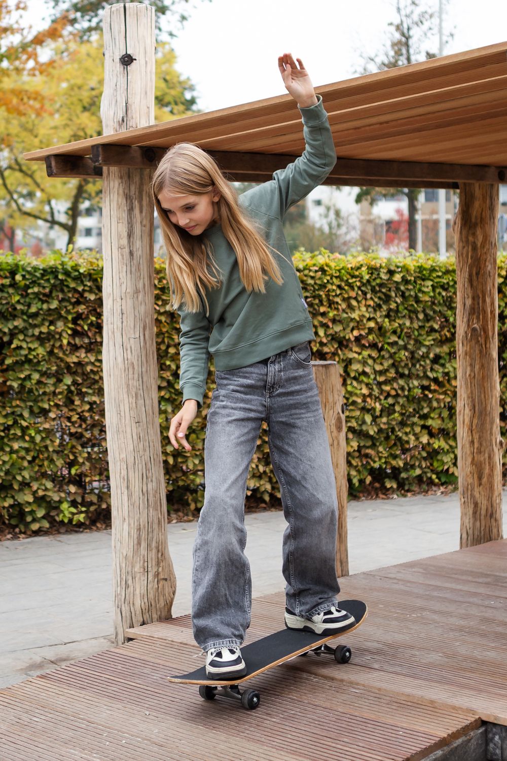 A skateboarder in jeans and green sweater practices balancing tricks on a wooden deck during autumn.
