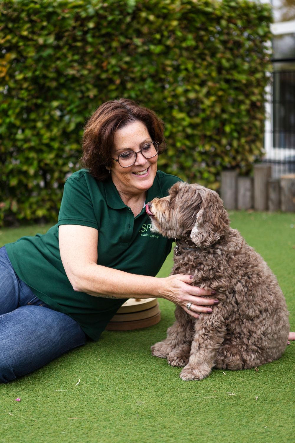 A person in a green shirt lying on grass while petting and playing with a small brown shaggy dog.