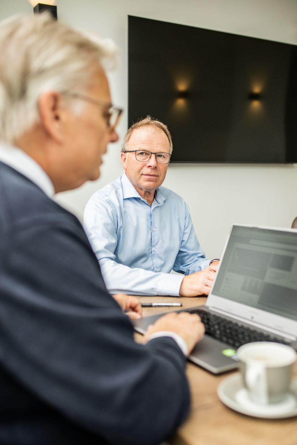 Two businessmen in a professional meeting, one listening attentively while the other works on a laptop.