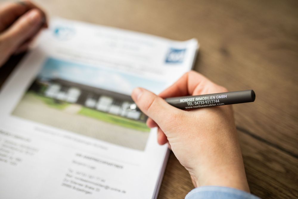 Close-up of a hand holding a pen over a real estate property brochure on a wooden desk.