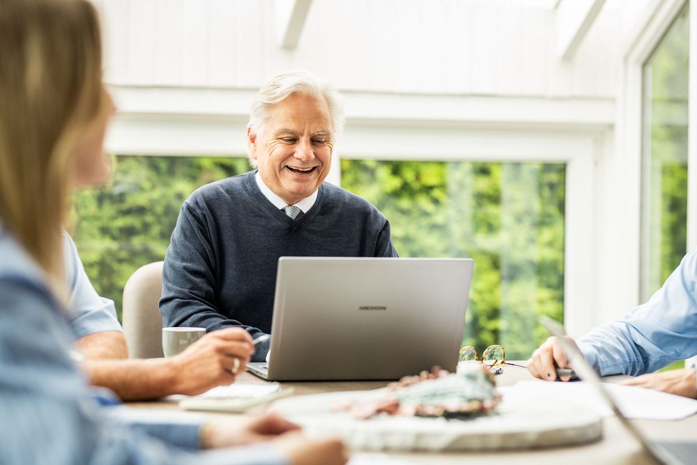 Smiling senior businessman showing laptop screen to colleagues during a meeting in a sunlit conservatory.