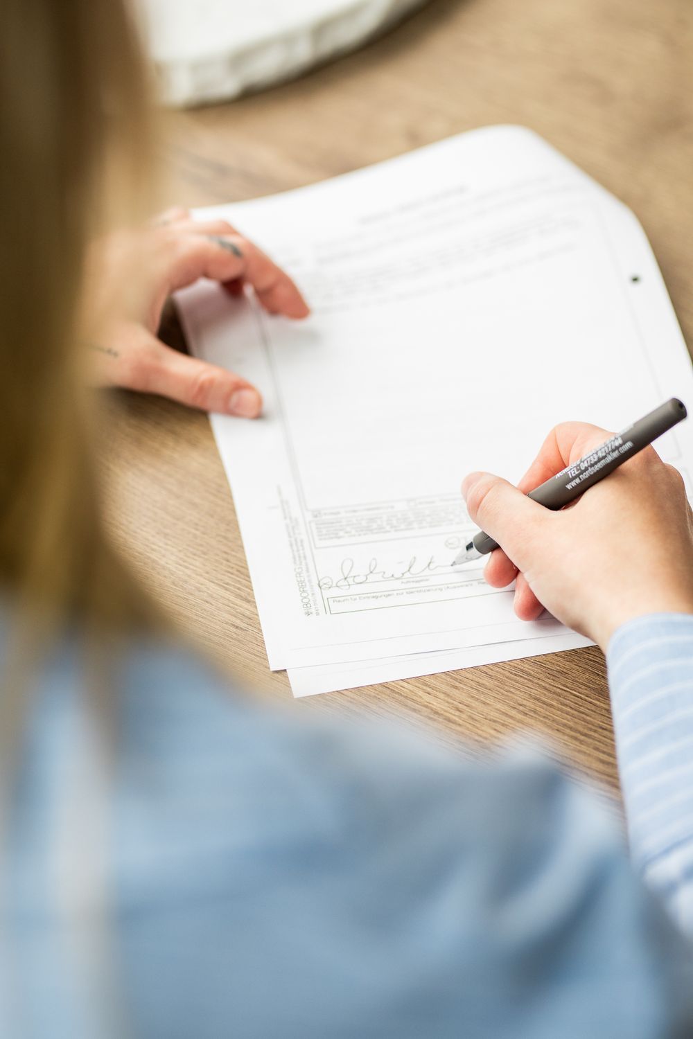 Close-up of a woman signing important documents with a black pen at a wooden desk.