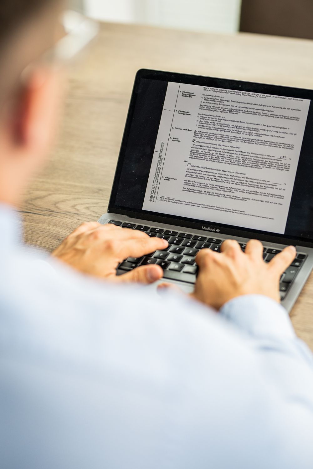 Businessman typing on a MacBook laptop displaying a formal document at a wooden desk.