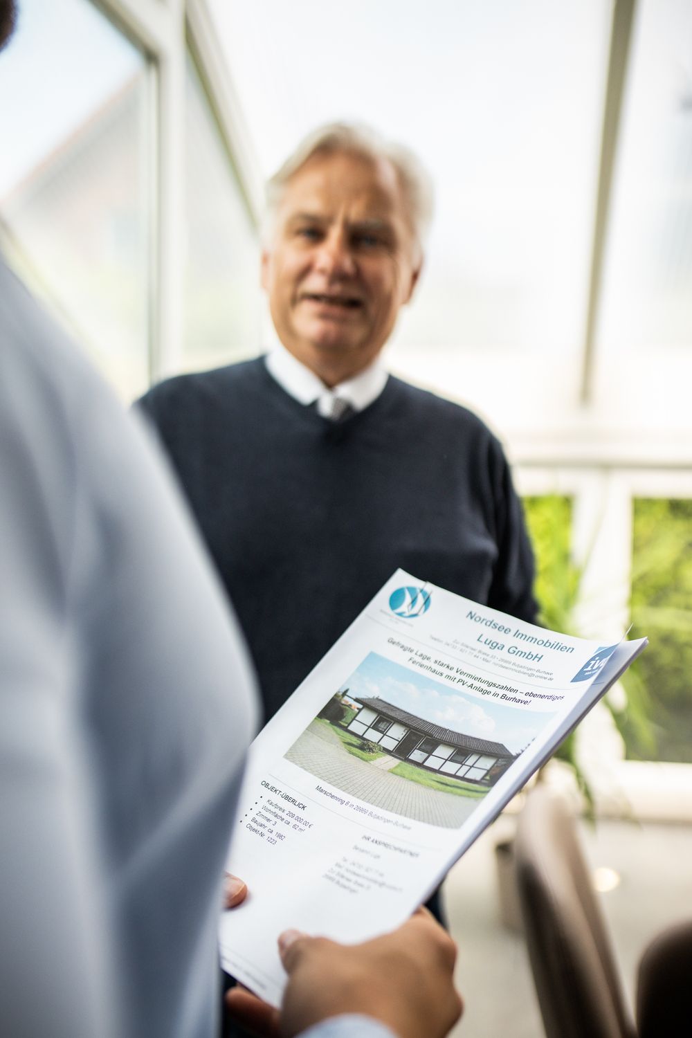 Senior businessman holding a real estate property brochure while speaking with a colleague indoors.