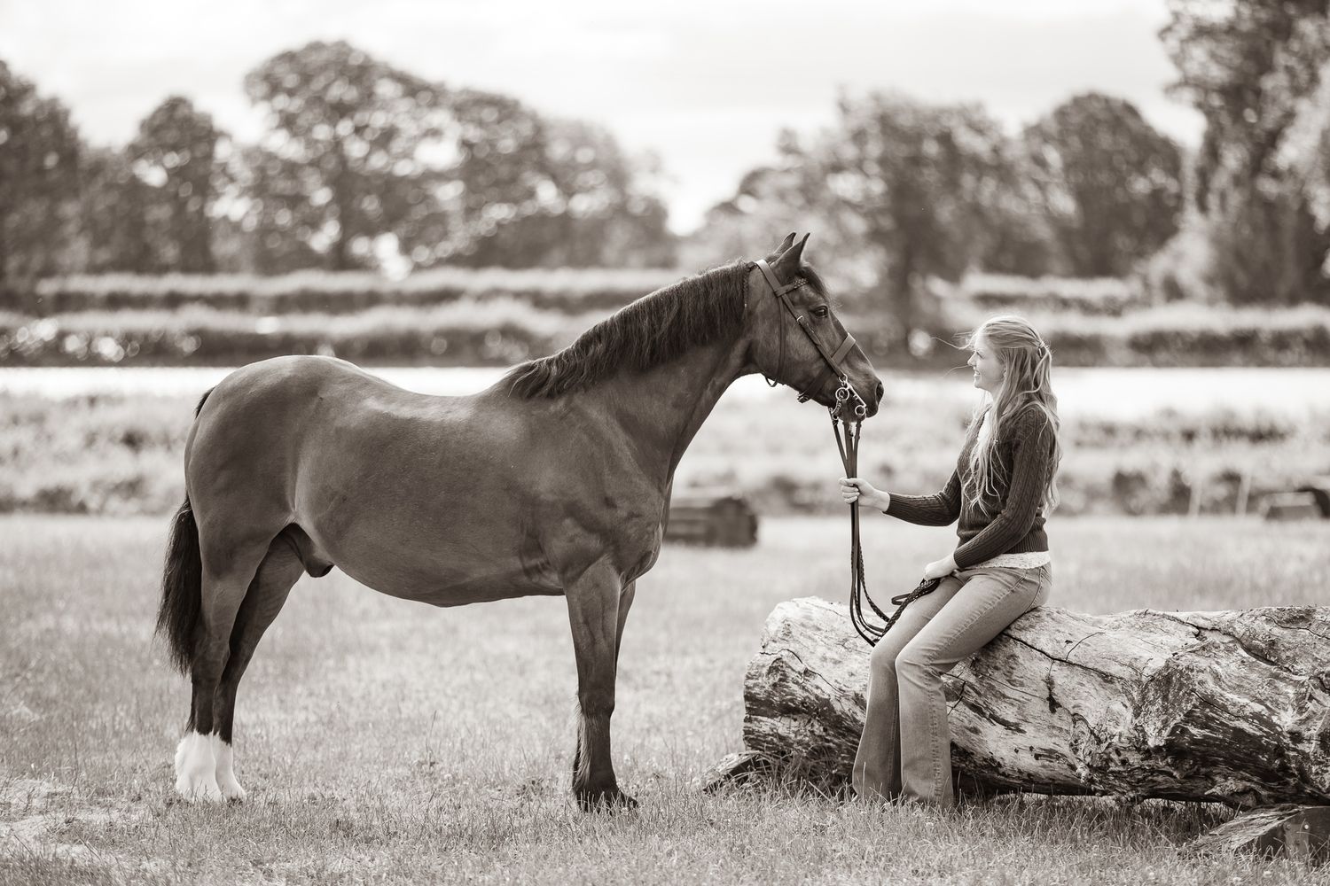 A woman sits on a log beside a brown horse in a field, holding its reins in a serene black and white photo.