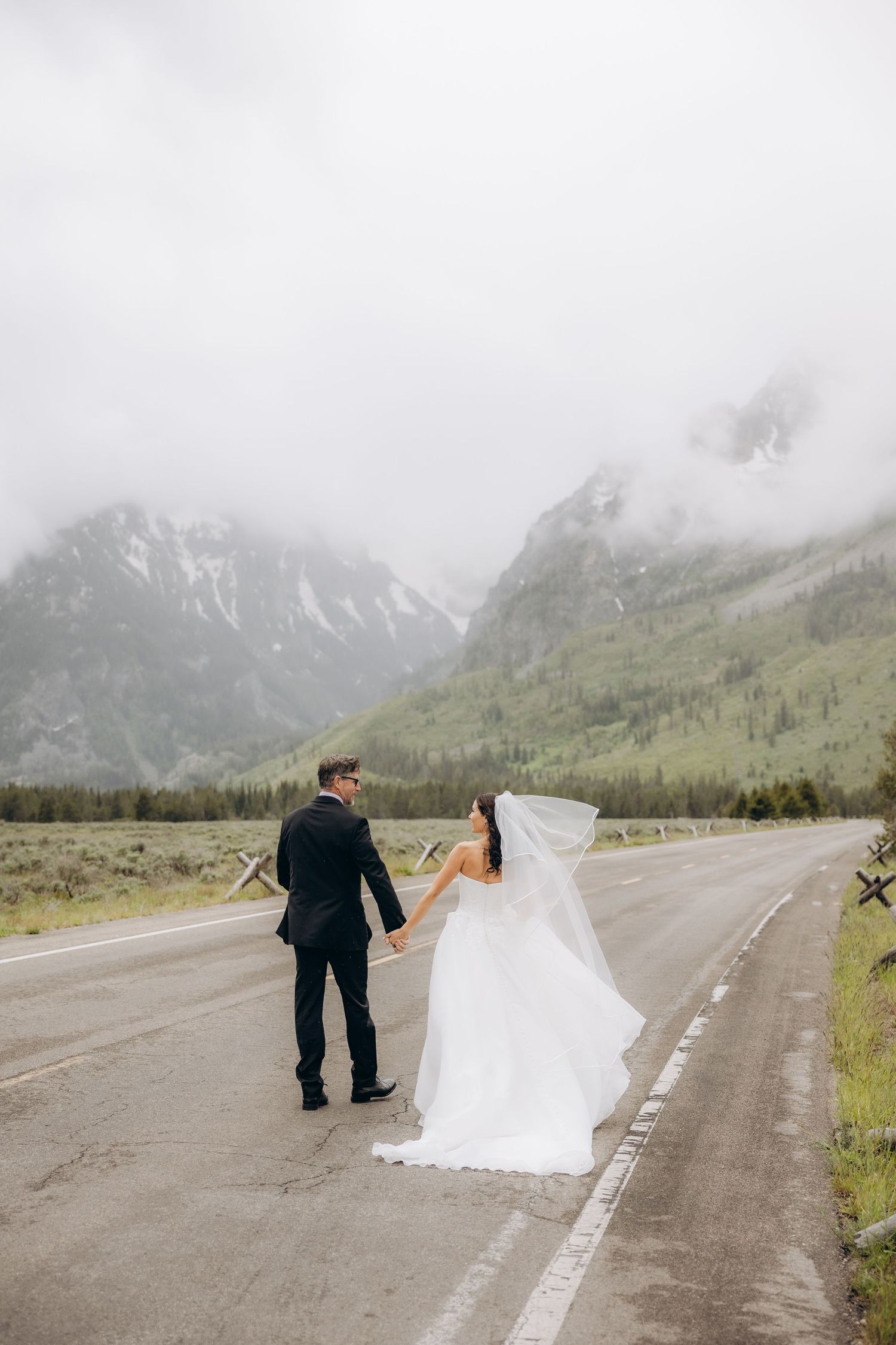 Bride and groom hold hands on a misty mountain road in Grand Teton, her veil flowing in the wind.