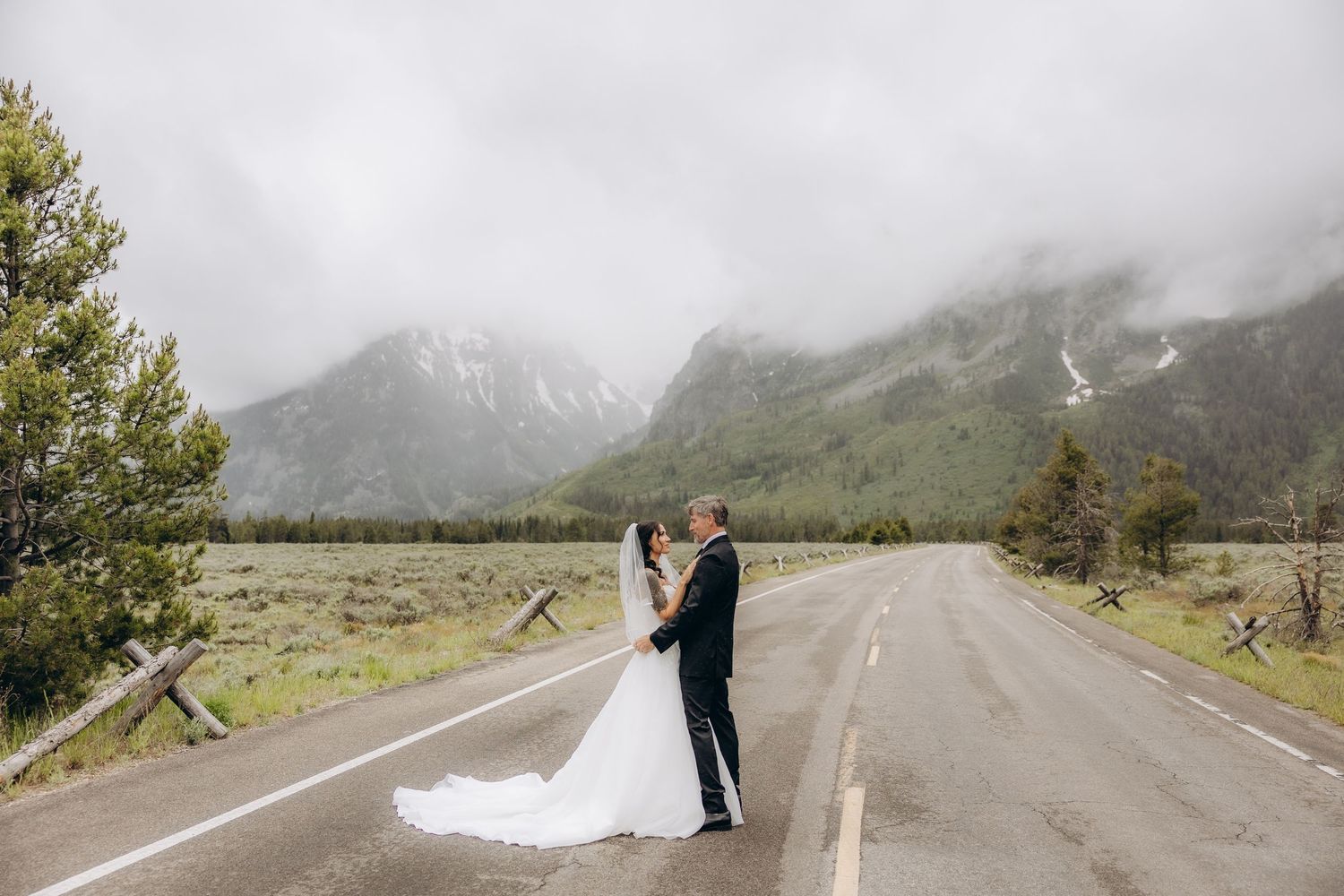 A bride and groom share a romantic kiss on an empty mountain road surrounded by misty peaks and lush greenery.