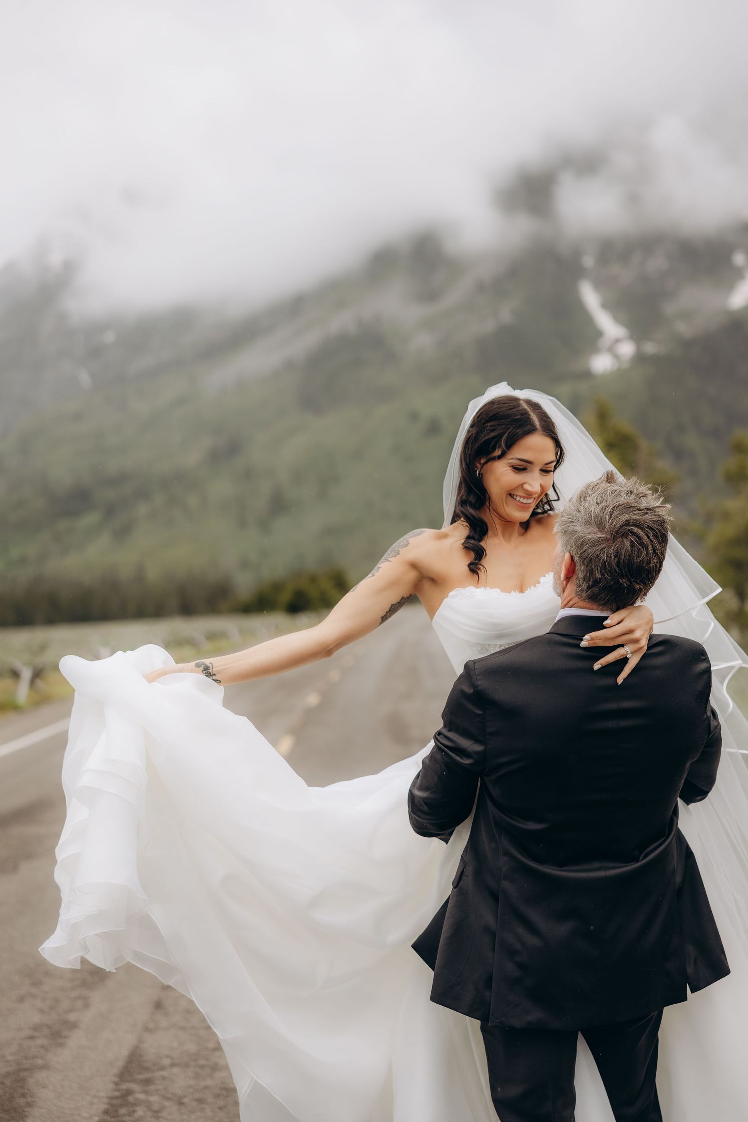 Bride in flowing white gown laughs as groom holds her on misty mountain road.