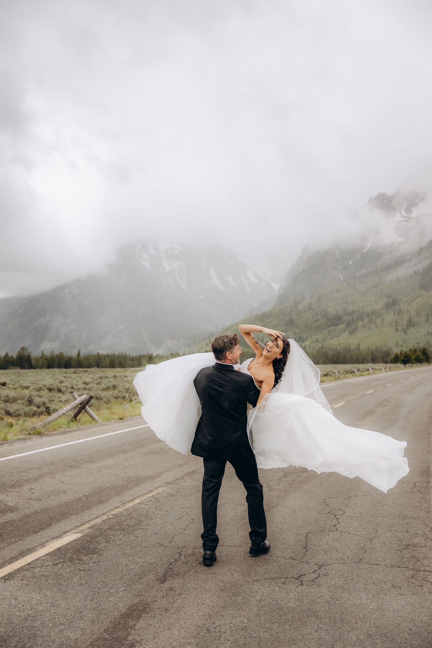 Bride and groom dancing on misty mountain road, white gown flowing in wind, dramatic cloudy peaks behind them.