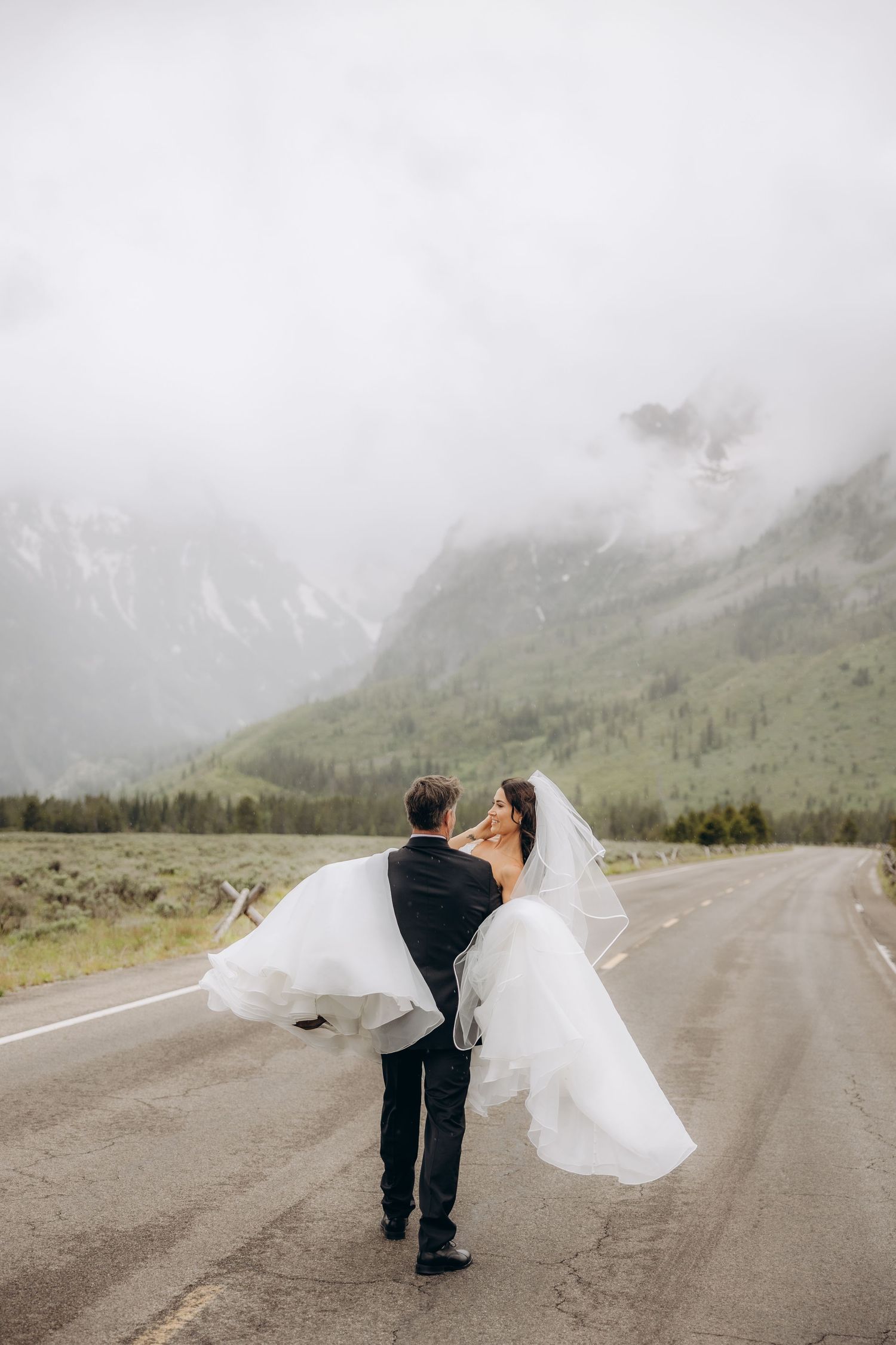 Groom lifts bride in white dress on foggy mountain highway during wedding shoot.