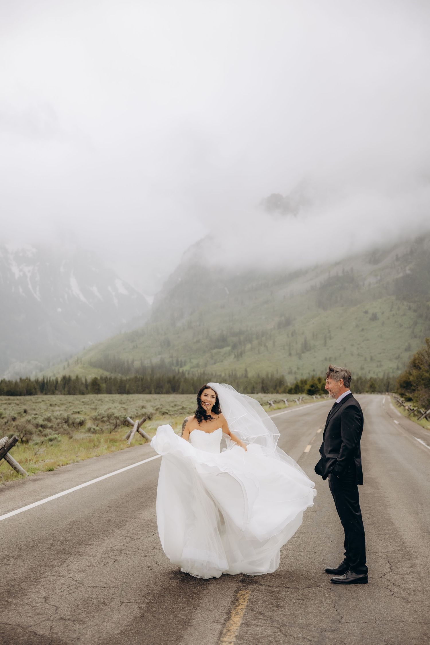 Bride in flowing white gown twirls on misty mountain road while groom watches in Grand Teton National Park.
