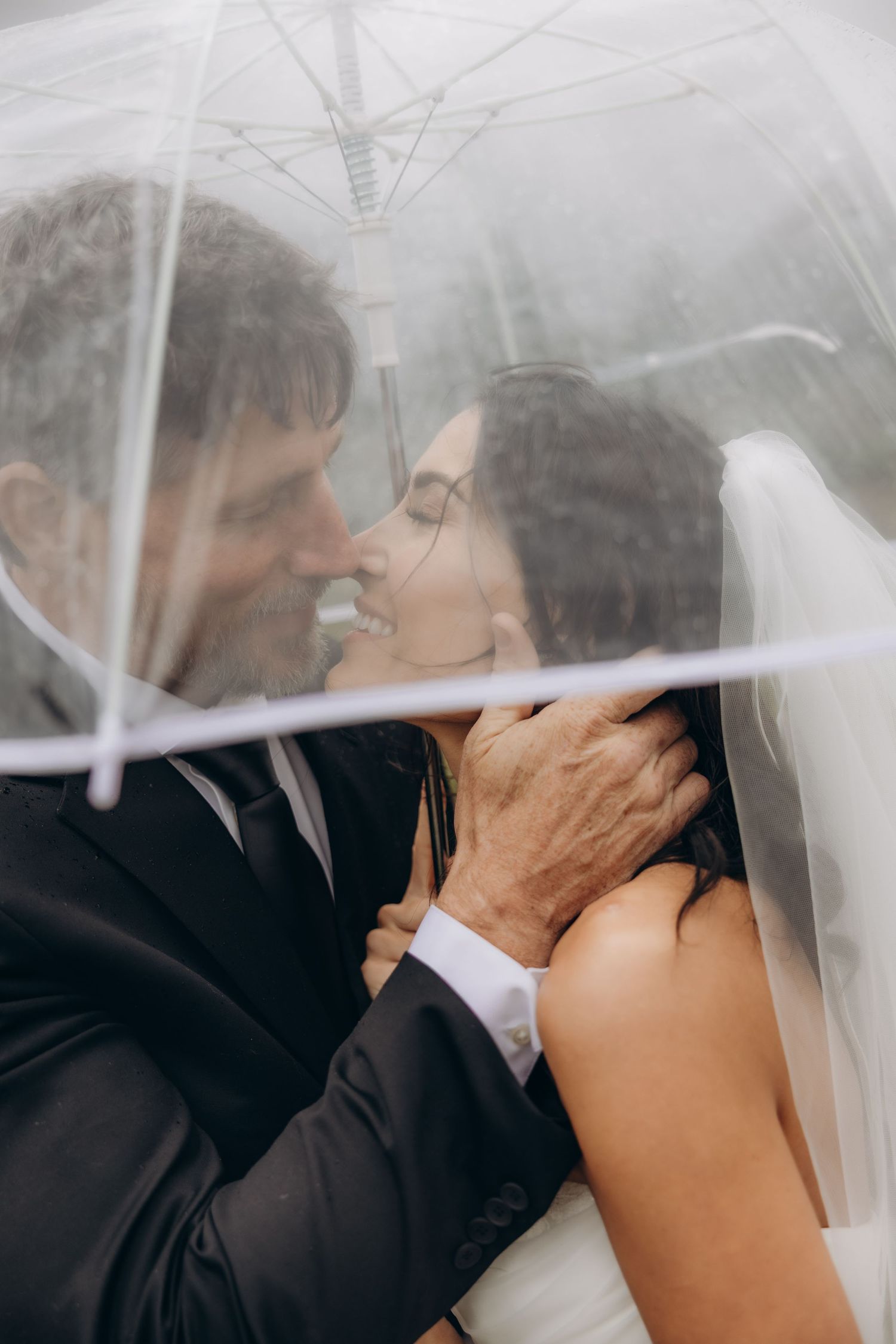 Bride and groom sharing romantic kiss under clear umbrella in rain, groom tenderly holding brides face.