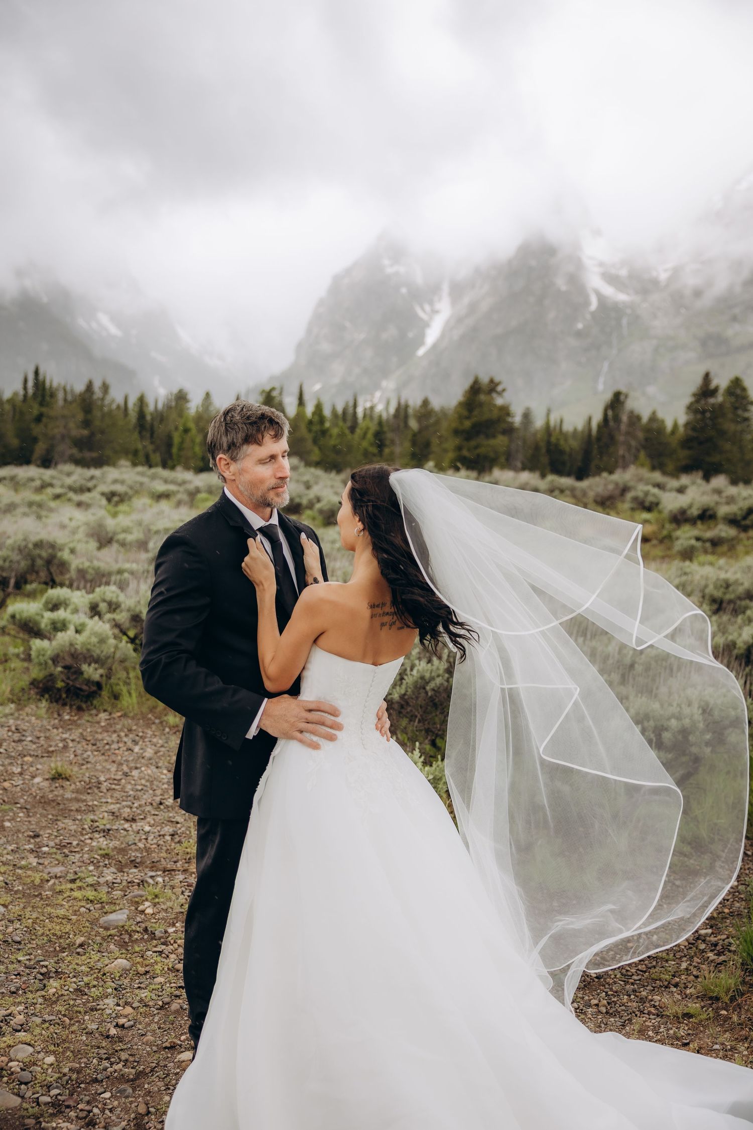 Bride and groom embrace in front of misty Grand Teton mountains, her veil flowing in the wind.
