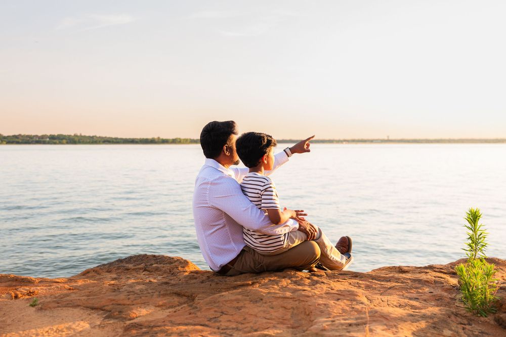 Father and child sitting on rocky shore pointing at scenic lake during golden sunset.