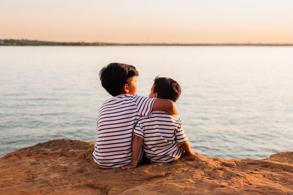 Two children in striped shirts sit together on a rock overlooking a calm lake at sunset.