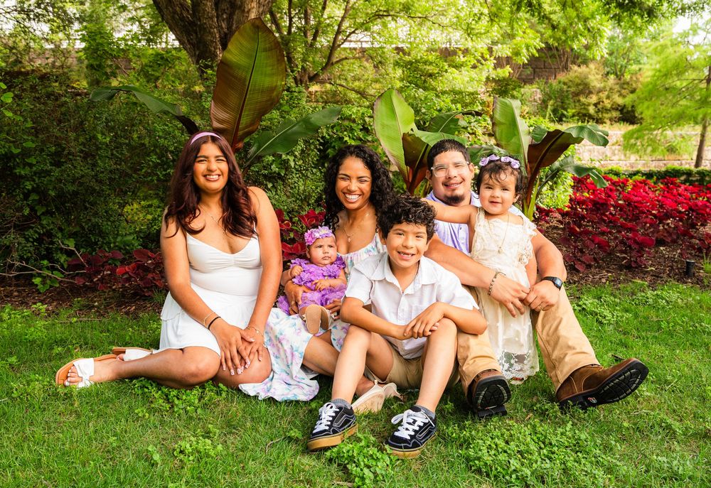 Happy family of six posing outdoors on green grass surrounded by lush garden trees and red flowers.