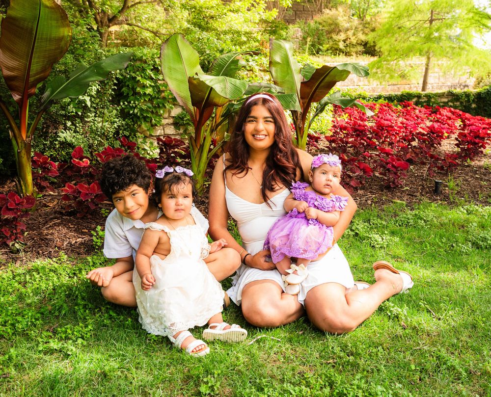 Woman in white dress sits on grass with three young children, surrounded by lush green and red garden foliage.