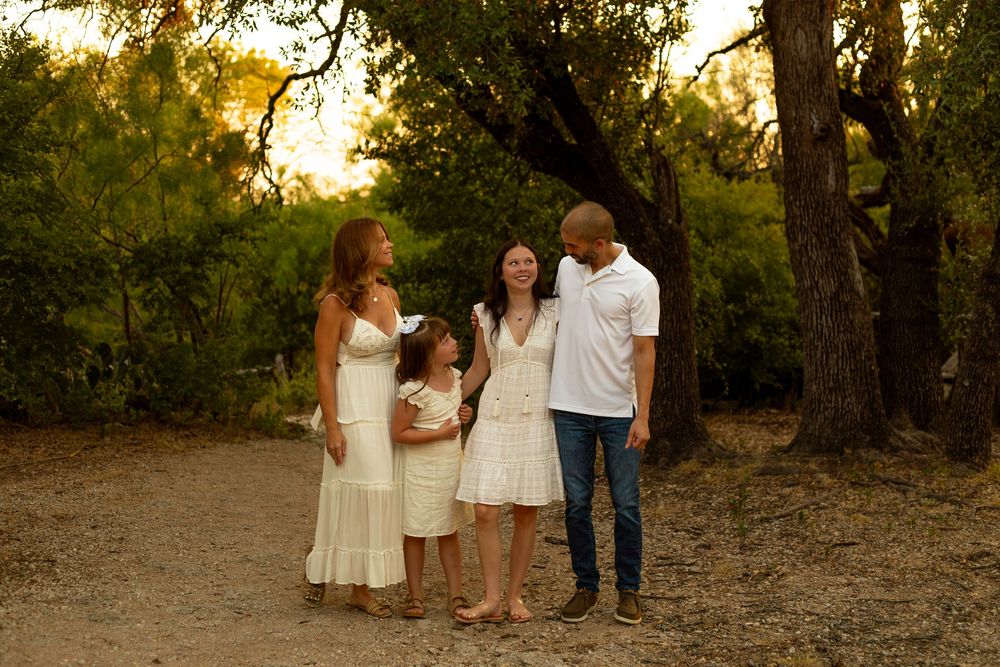 Family of four in white outfits posing outdoors among lush green trees during golden hour sunset.