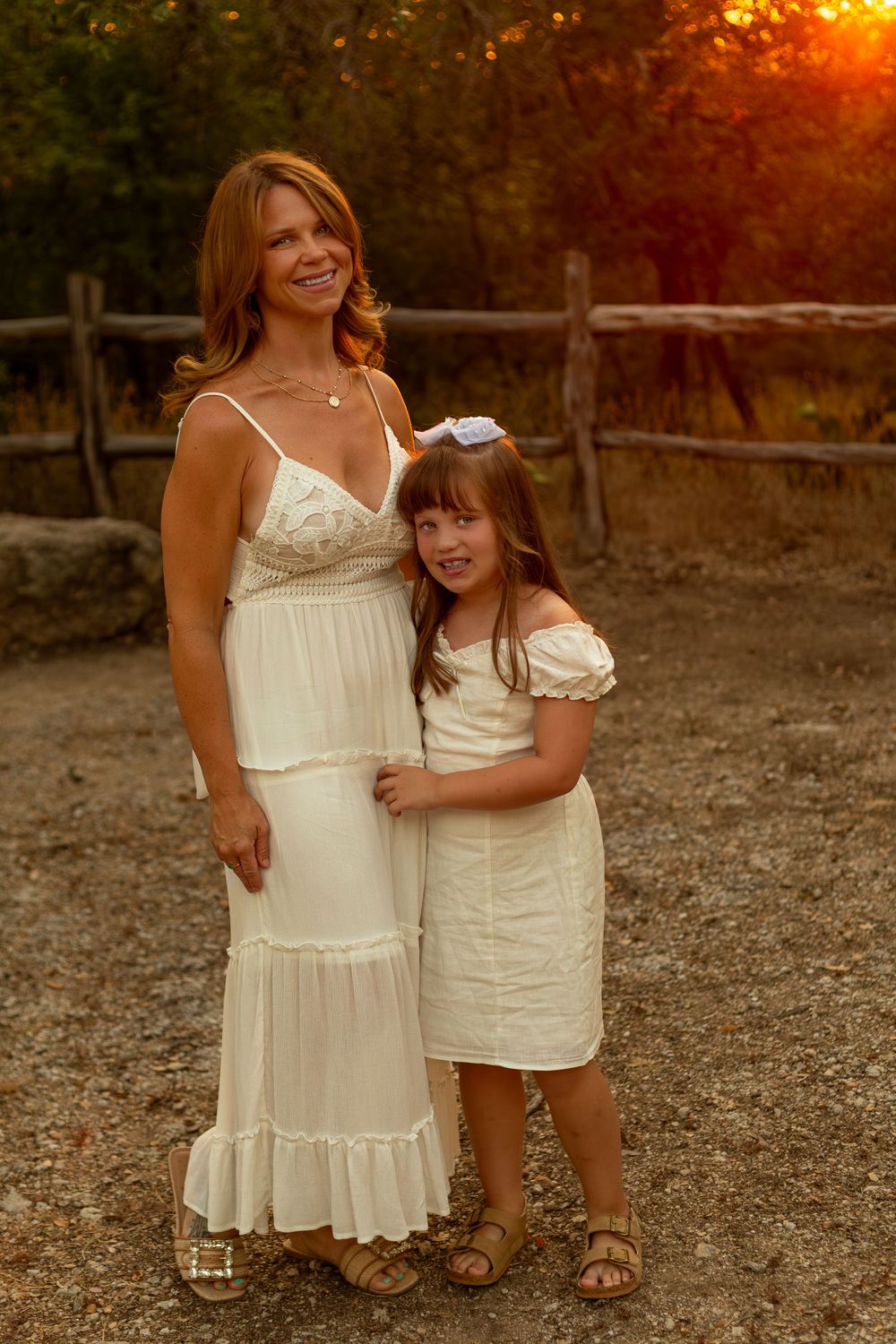 Mother and daughter in matching white dresses pose together outdoors at golden hour near a rustic wooden fence.