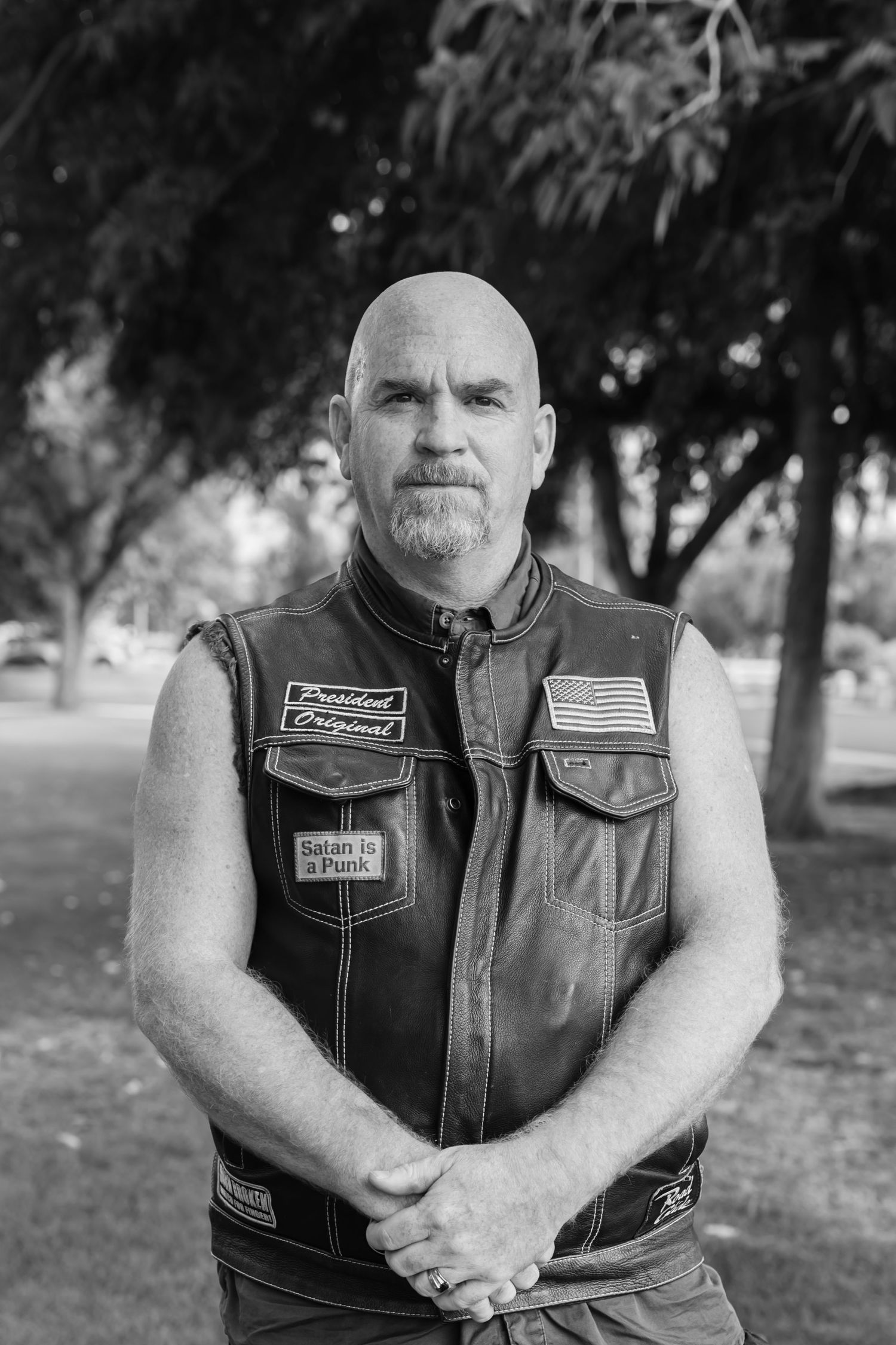 Bald man with goatee wearing a leather biker vest with patches stands outdoors in black and white photo.