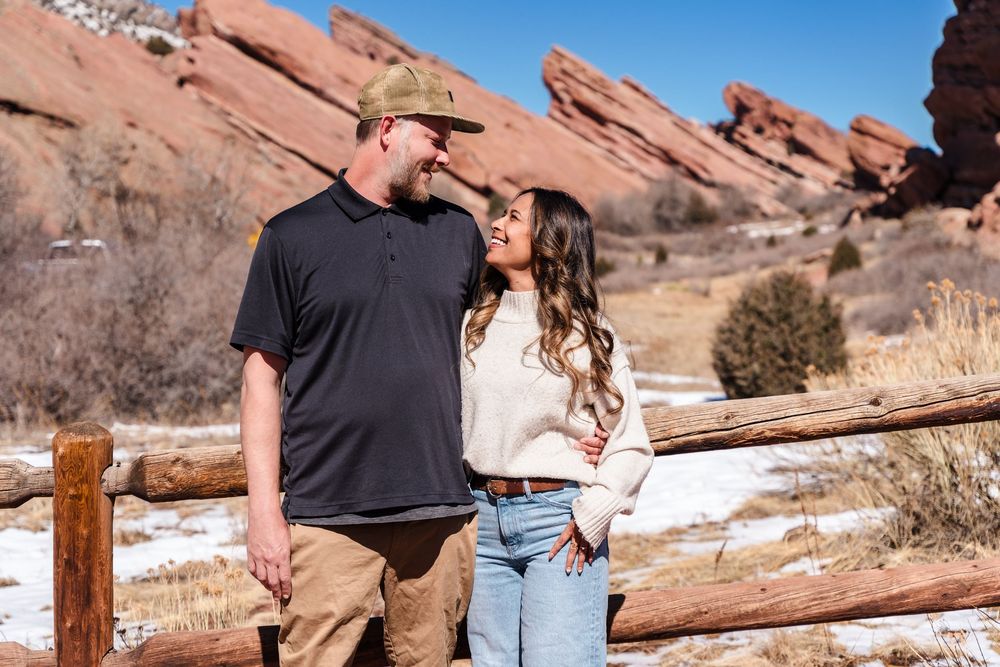 Couple smiling at each other at Red Rocks Park Colorado with red rock formations and snow in background.