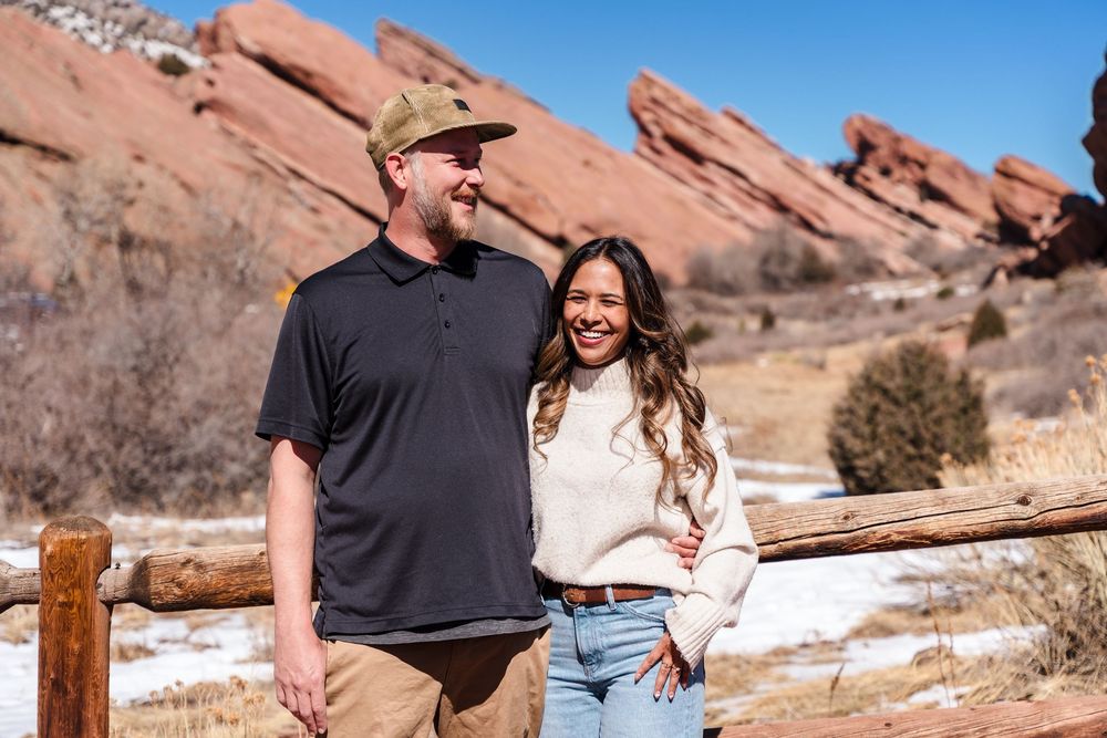 Couple smiling together at Red Rocks Park in Colorado with iconic red rock formations and snow in the background.