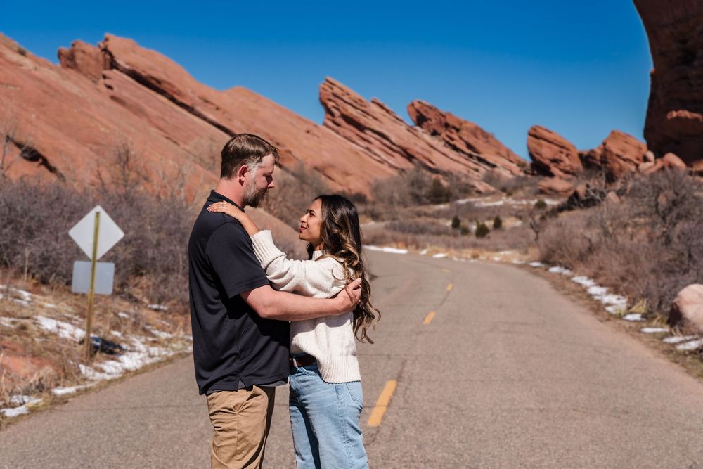Couple embracing on a road at Red Rocks Park, Colorado, with iconic red sandstone formations under a clear blue sky.