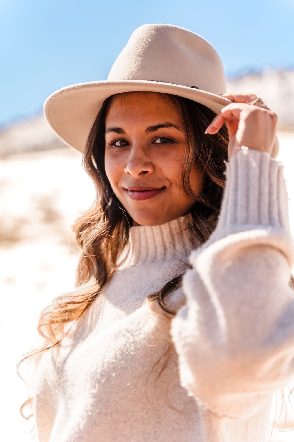 A young woman in a cream turtleneck sweater and beige wide-brim hat smiles outdoors in a sunny desert setting.